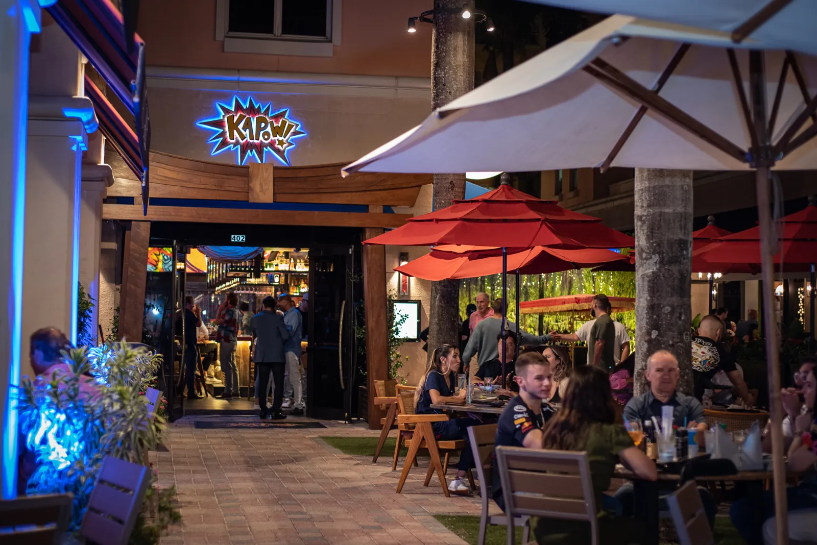 Evening outdoor dining at a restaurant with neon 'KAPOW!' sign and red patio umbrellas