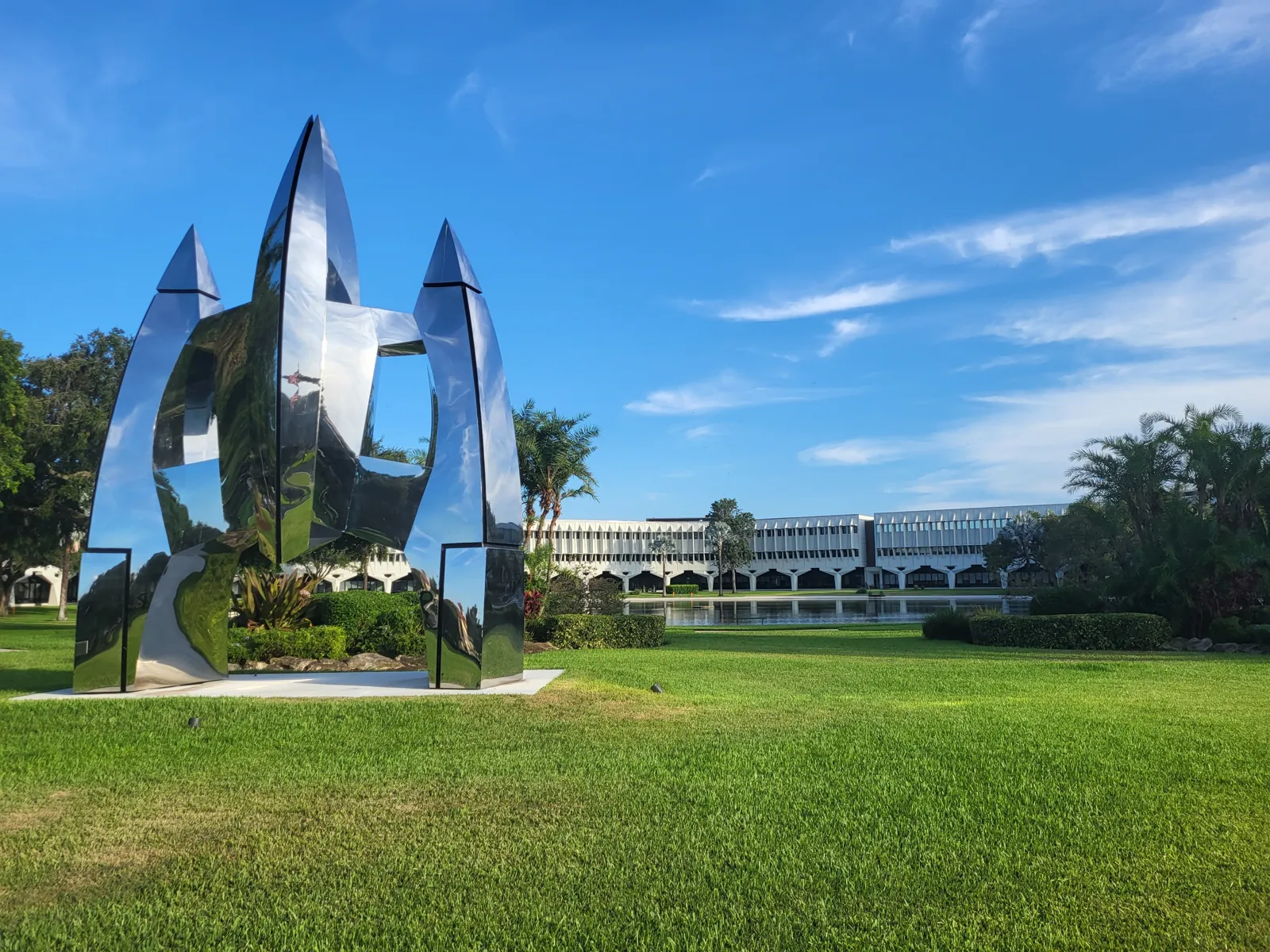 Polished stainless-steel abstract sculpture on manicured lawn with modernist campus building and reflecting pond
