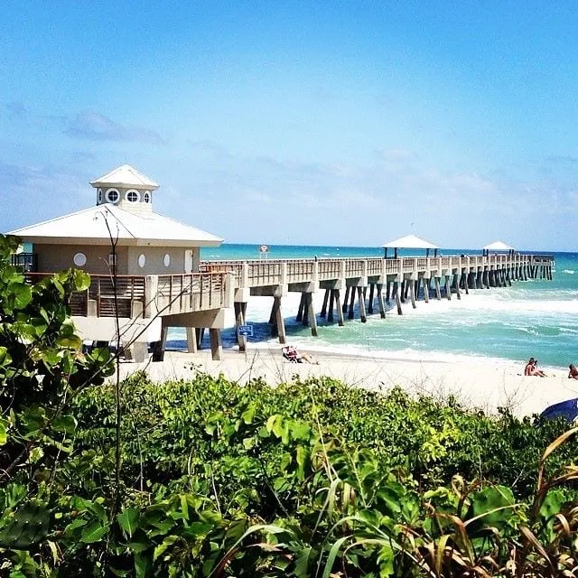 The Juno Beach Pier and its pier house, seen over dune plants, with sunbathers on the sand and turquoise water beyond