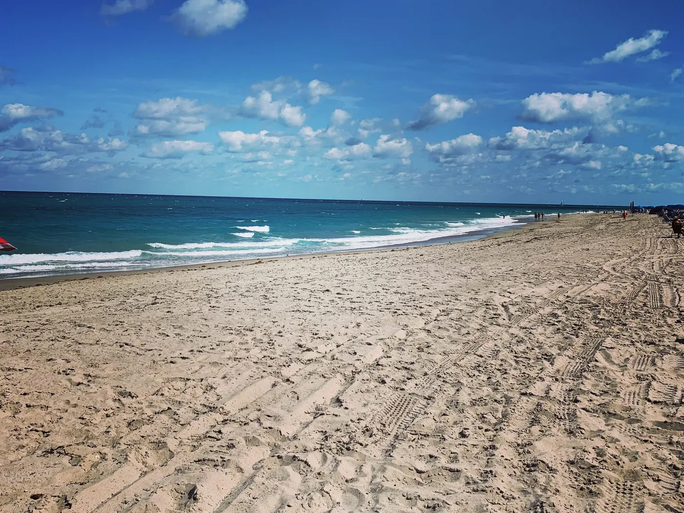 A wide sandy beach in Juno Beach with turquoise surf and a few distant beachgoers under a blue sky with puffy clouds