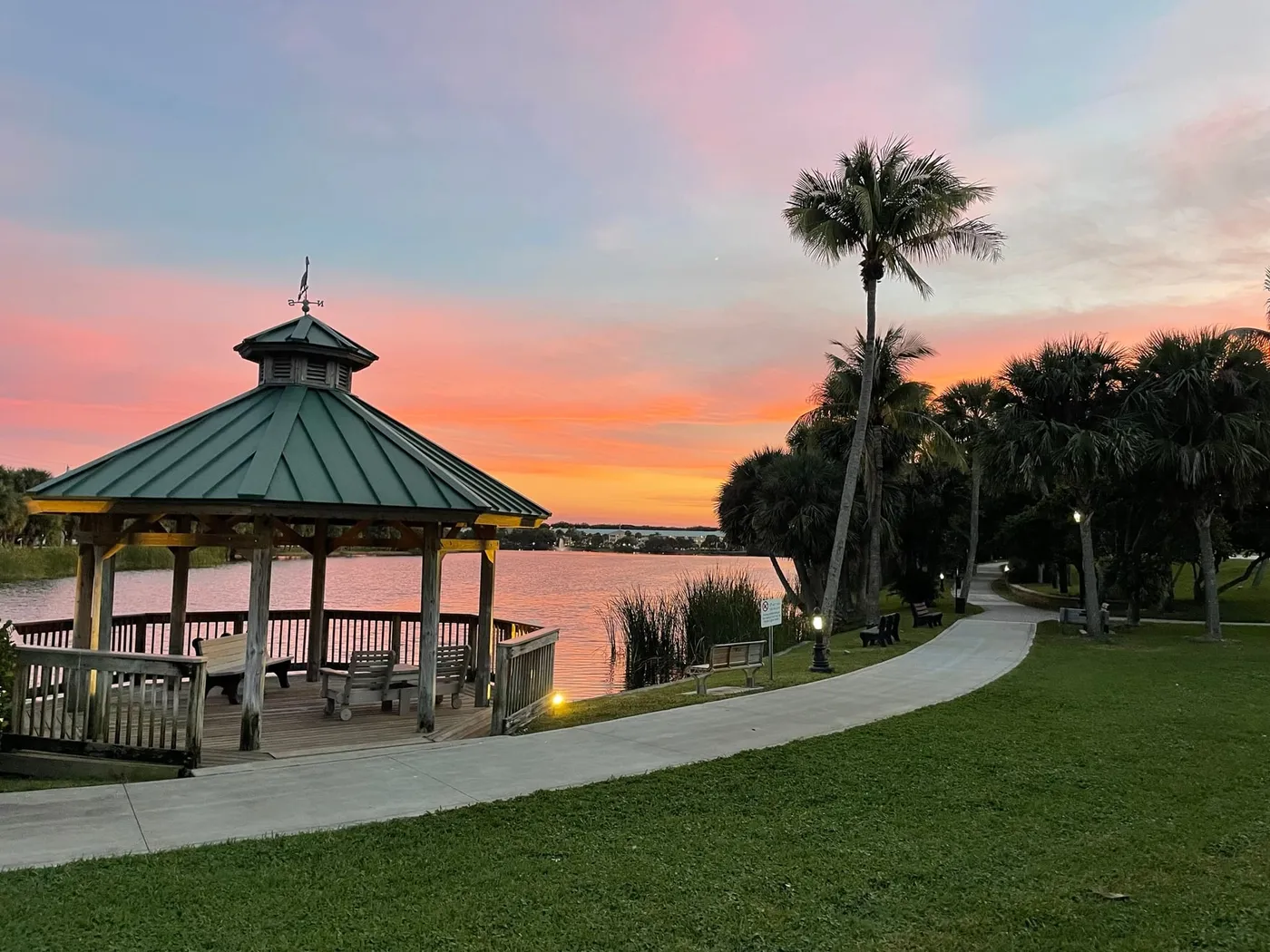 A lakeside gazebo with a heron weathervane at sunset, with a curving path, benches and palms