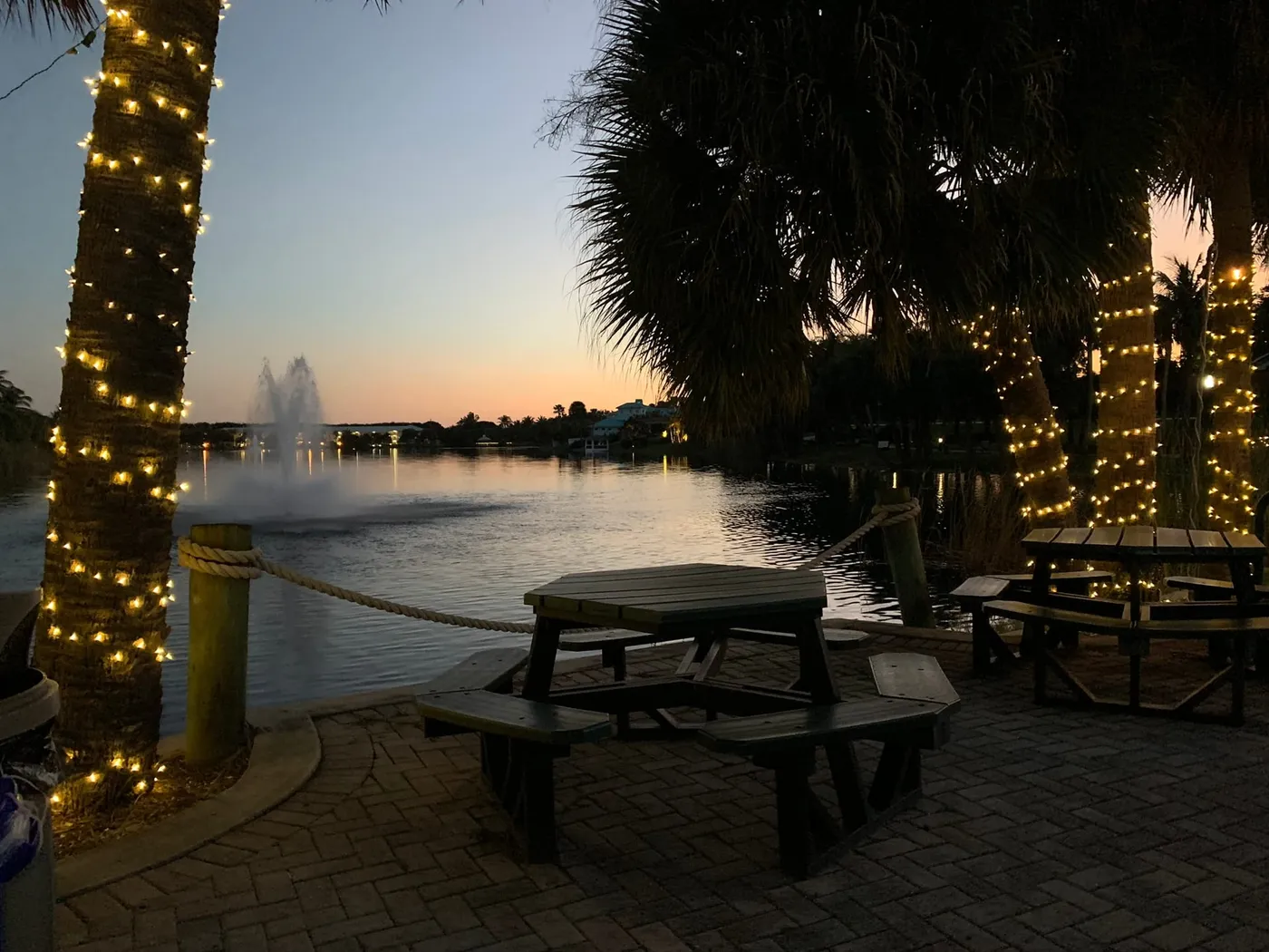 A brick patio with octagonal picnic tables and string-lit palms beside Pelican Lake at dusk, with a fountain on the water