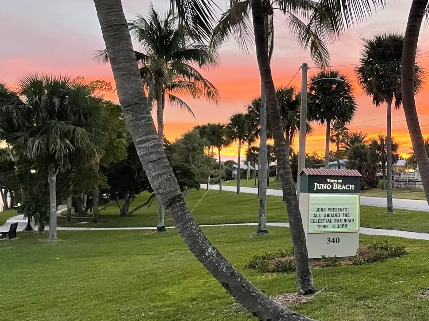 The Town of Juno Beach sign among palms at sunset, its marquee advertising a talk on the Celestial Railroad