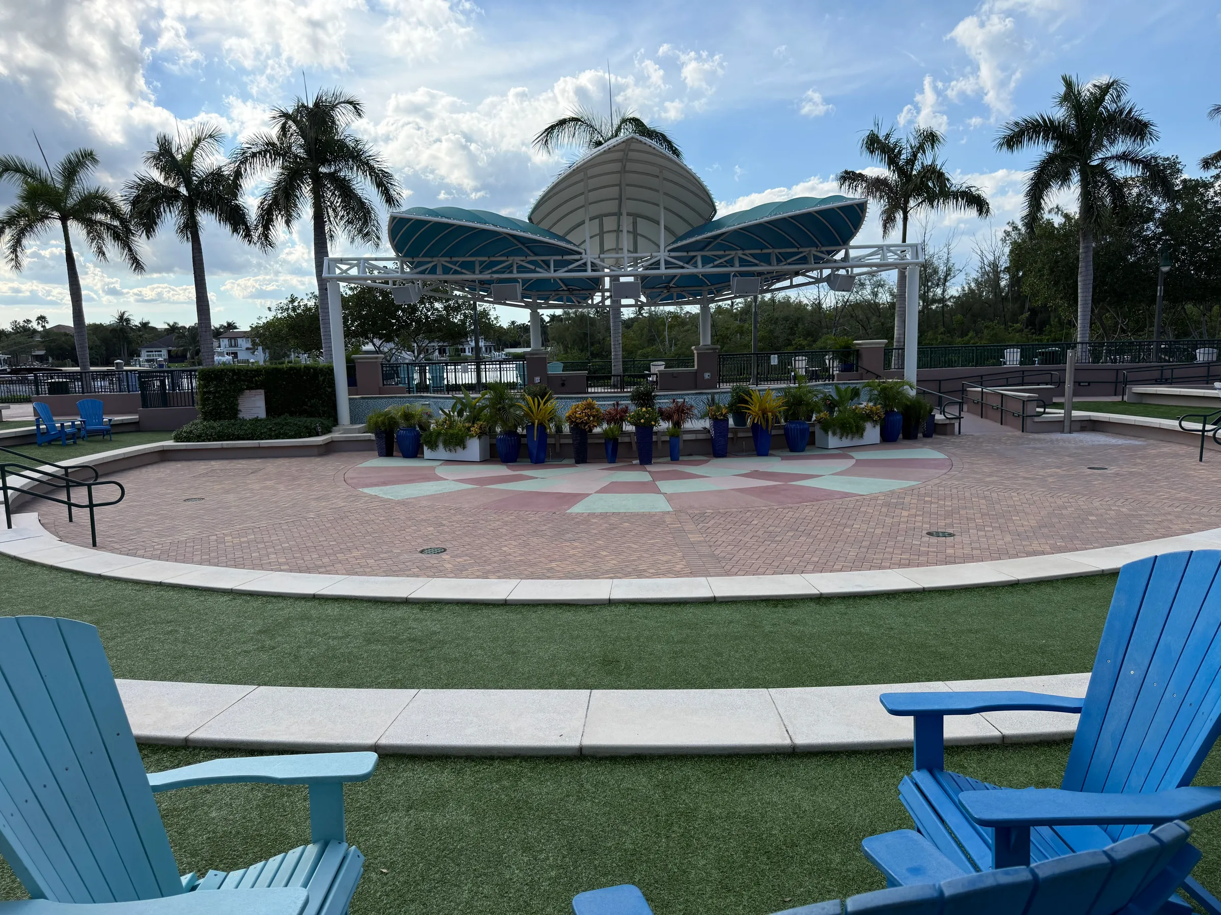 The Harbourside Place amphitheater stage under its teal canopy, with a lawn and blue Adirondack chairs in front and palms behind