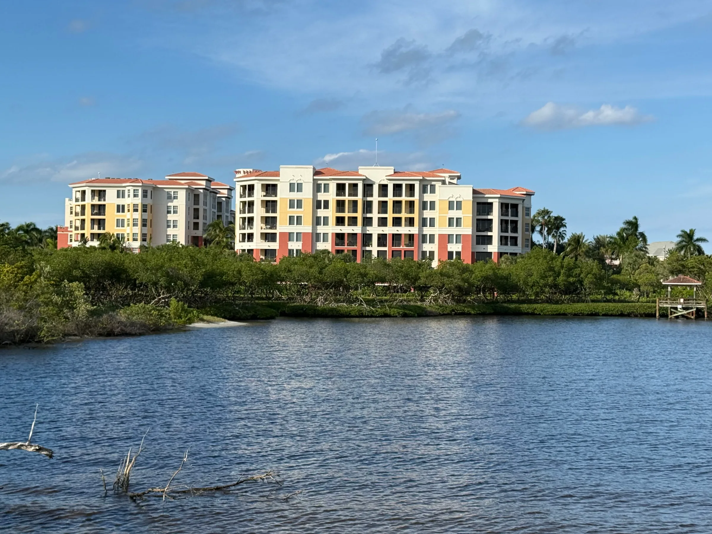 Two mid-rise condominium buildings in yellow and cream with terracotta roofs, seen across the water beyond a line of mangroves