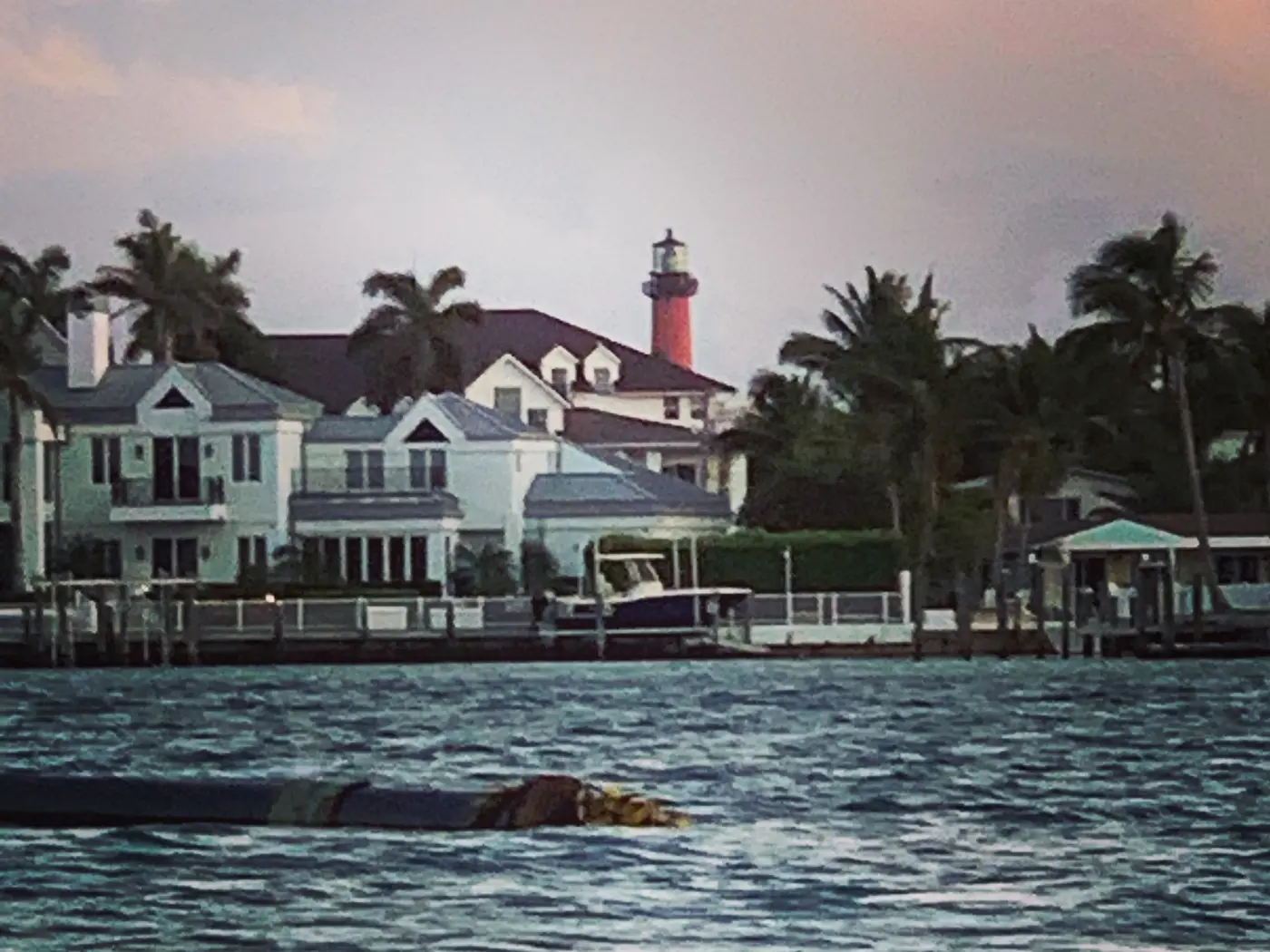 Two-story houses with docks and a moored boat on the water in Jupiter at sunset, with the Jupiter Inlet Lighthouse behind the rooftops