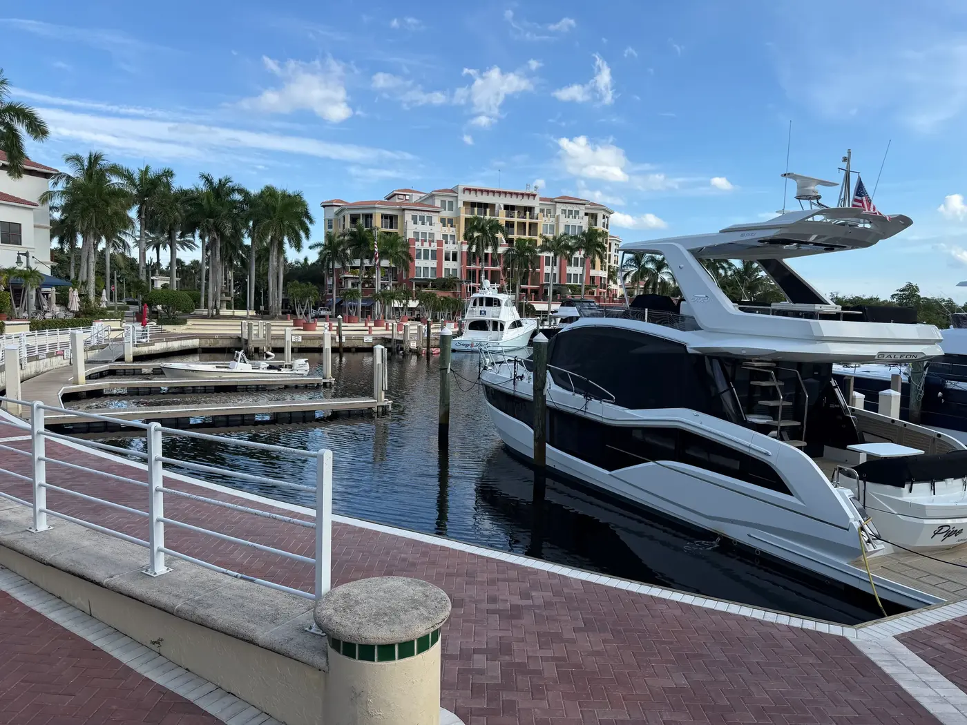 A brick promenade with white railings along a marina basin on the Jupiter Riverwalk, with a yacht in the foreground and condominium buildings behind
