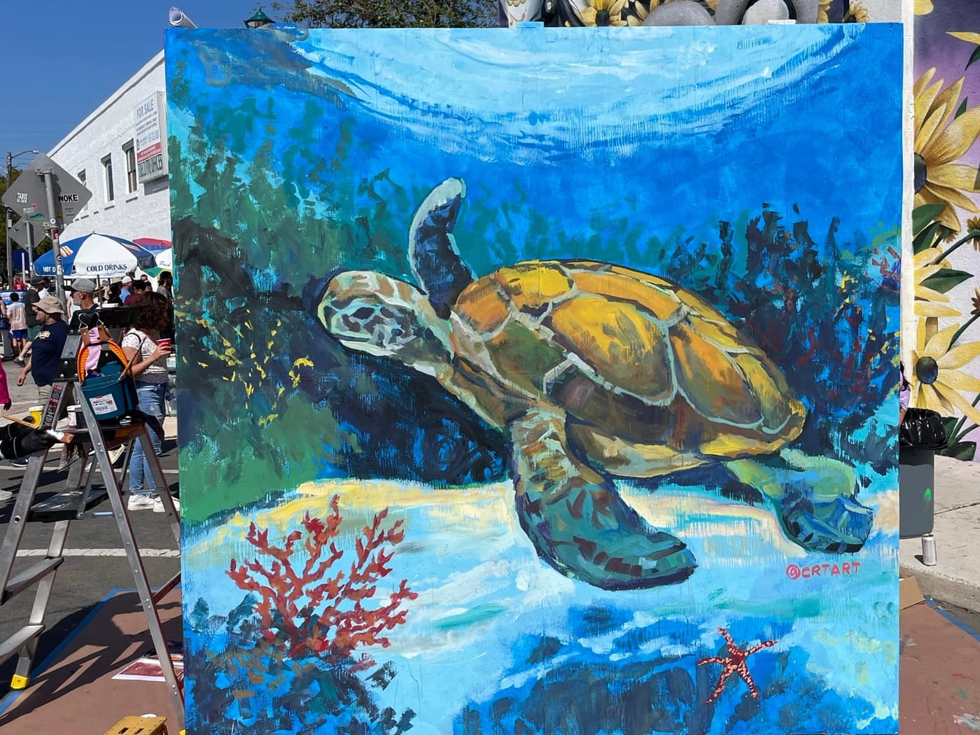A painted sea turtle panel on an easel at the Street Painting Festival, with a mural on the building behind it
