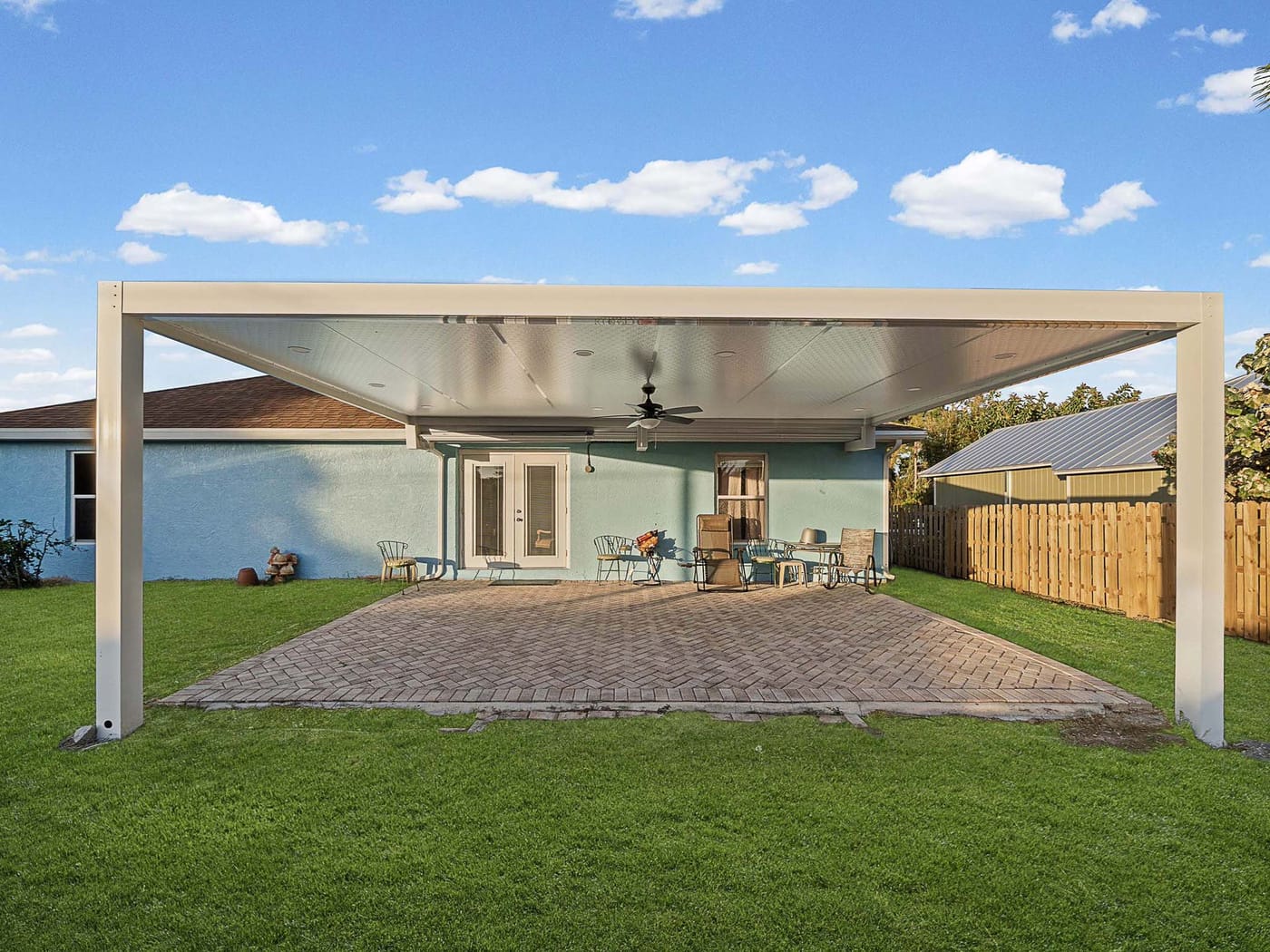A fenced backyard lawn with a white metal-roofed patio cover over a paver patio behind a single-story home
