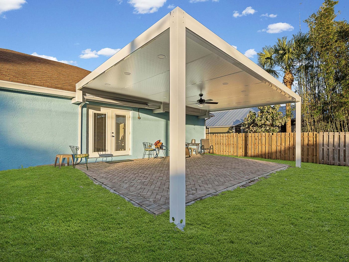 A white metal patio cover with a ceiling fan over a paver patio in a fenced backyard