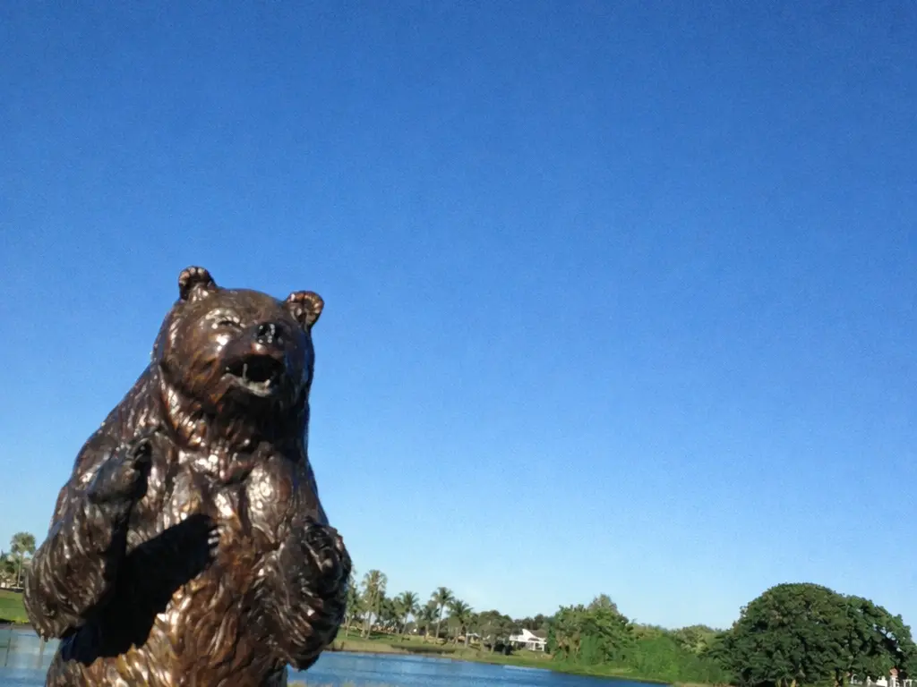 A bronze statue of a standing bear beside a lake and fairway on the Champion Course at PGA National in Palm Beach Gardens under a clear blue sky