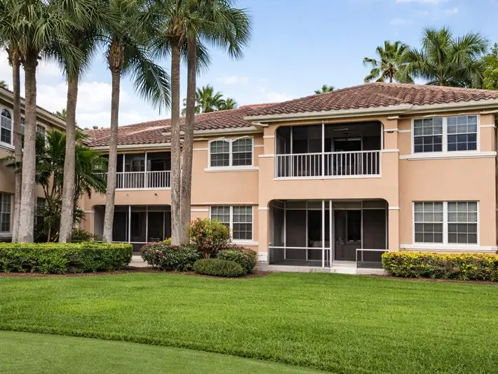 A two-storey condominium building with a barrel-tile roof and screened balconies behind a row of palms and a lawn at PGA National in Palm Beach Gardens