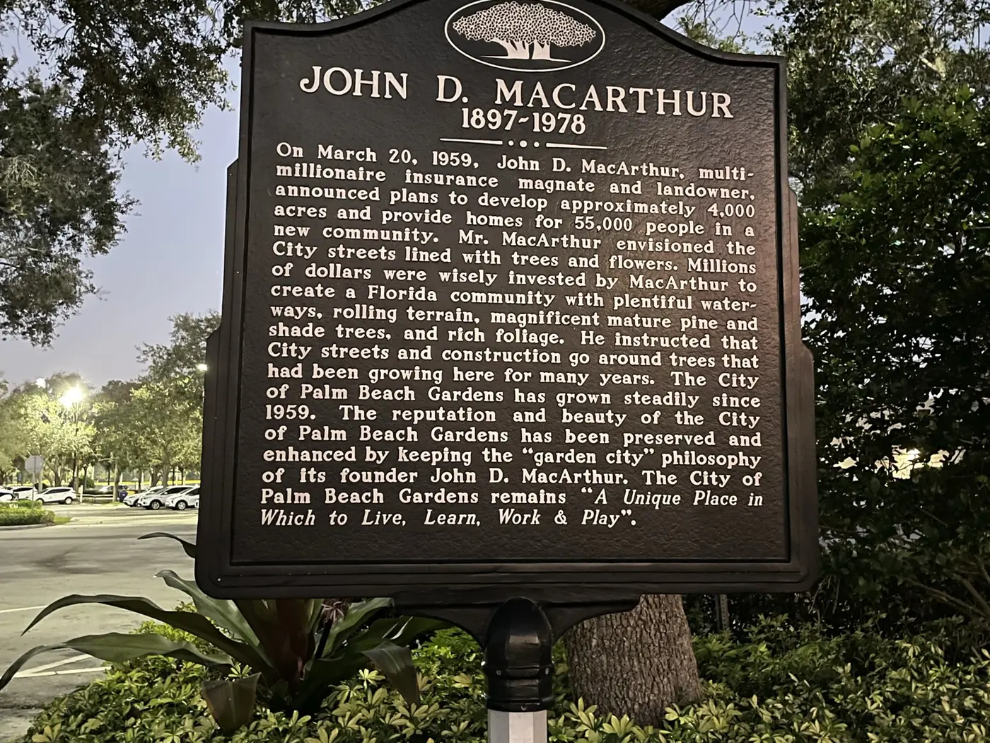 A black cast-metal historical marker headed John D. MacArthur 1897 to 1978 under a live oak at dusk in Palm Beach Gardens, its inscription describing the March 20, 1959 announcement of a planned community of approximately 4,000 acres