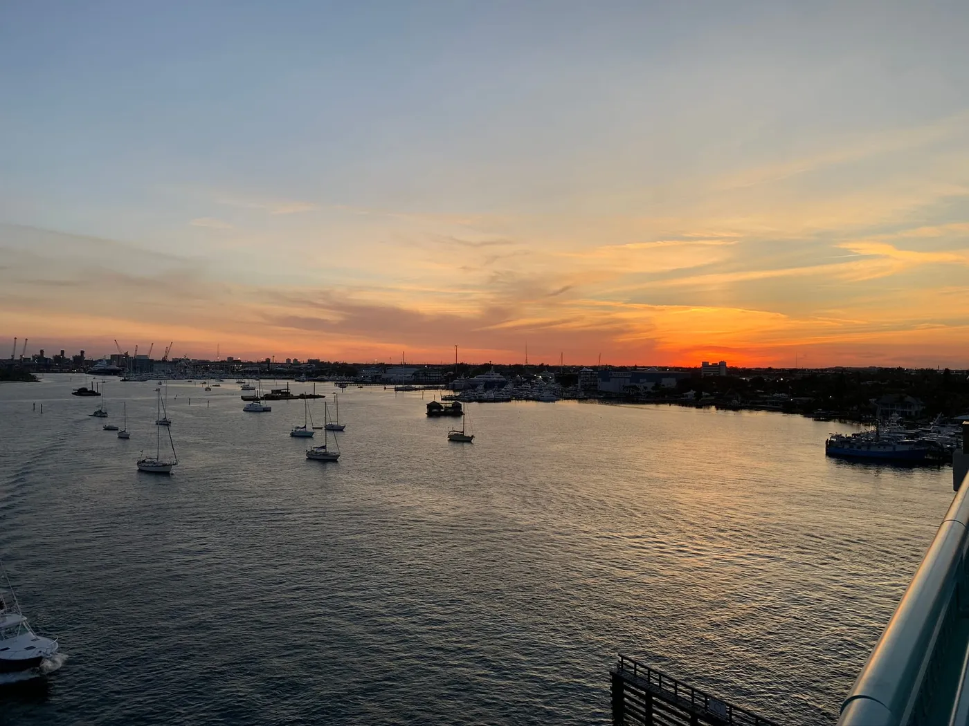 Sailboats at anchor in the lagoon at sunset under an orange sky, seen from the Blue Heron Bridge, with port cranes on the far horizon