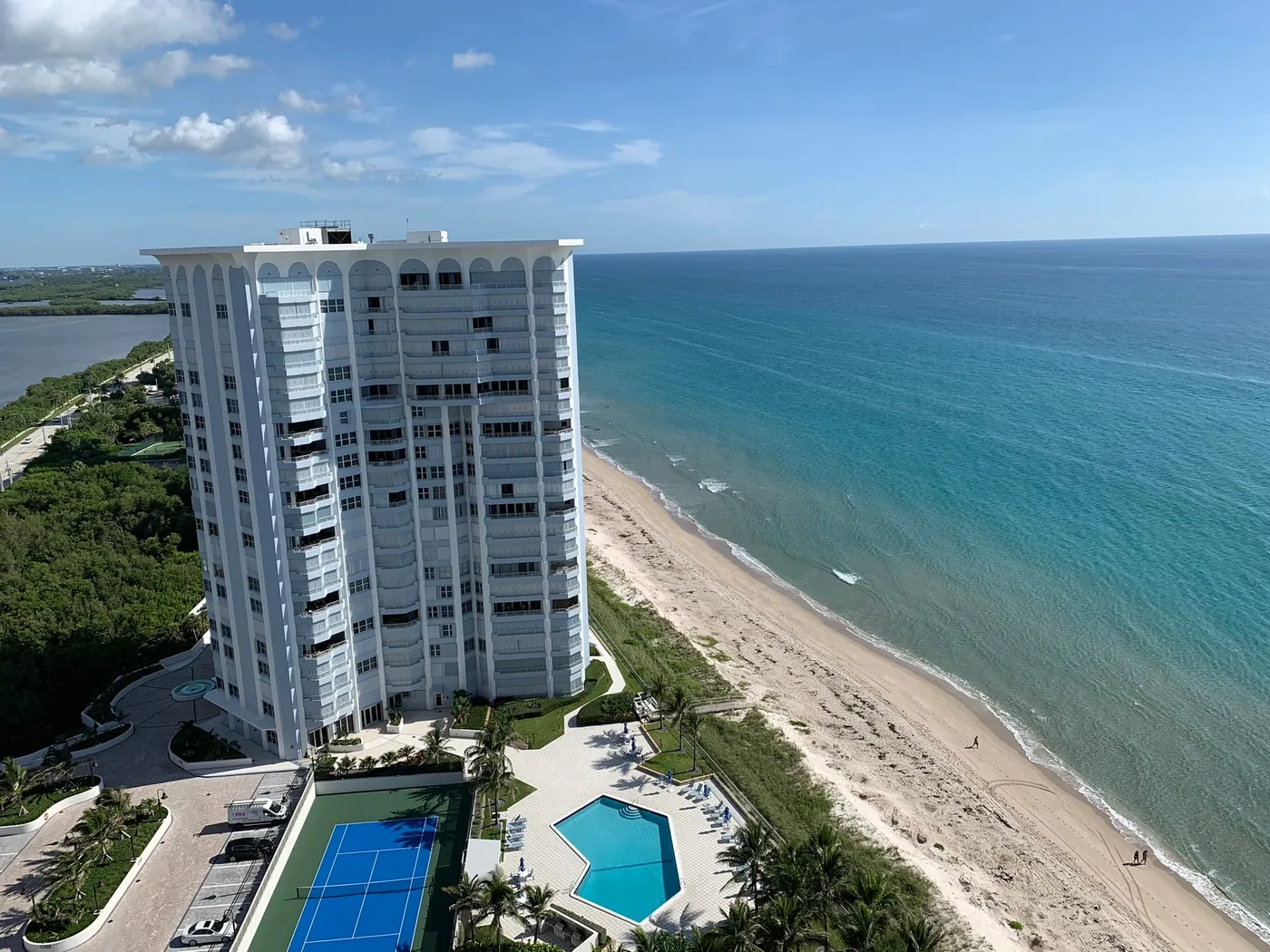 A white condominium tower on Singer Island beside the beach, with its tennis court and pool below and the lagoon and mangroves to the left