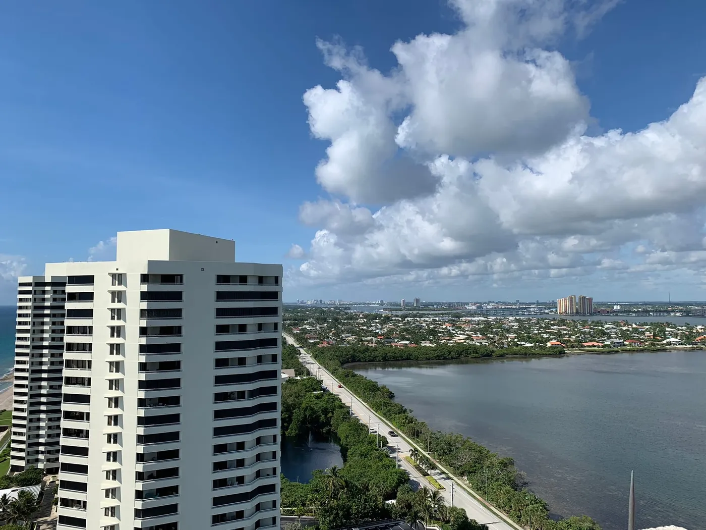 A white condominium tower beside the shore road on Singer Island, with the lagoon, the Blue Heron Bridge and the Port of Palm Beach cranes in the distance