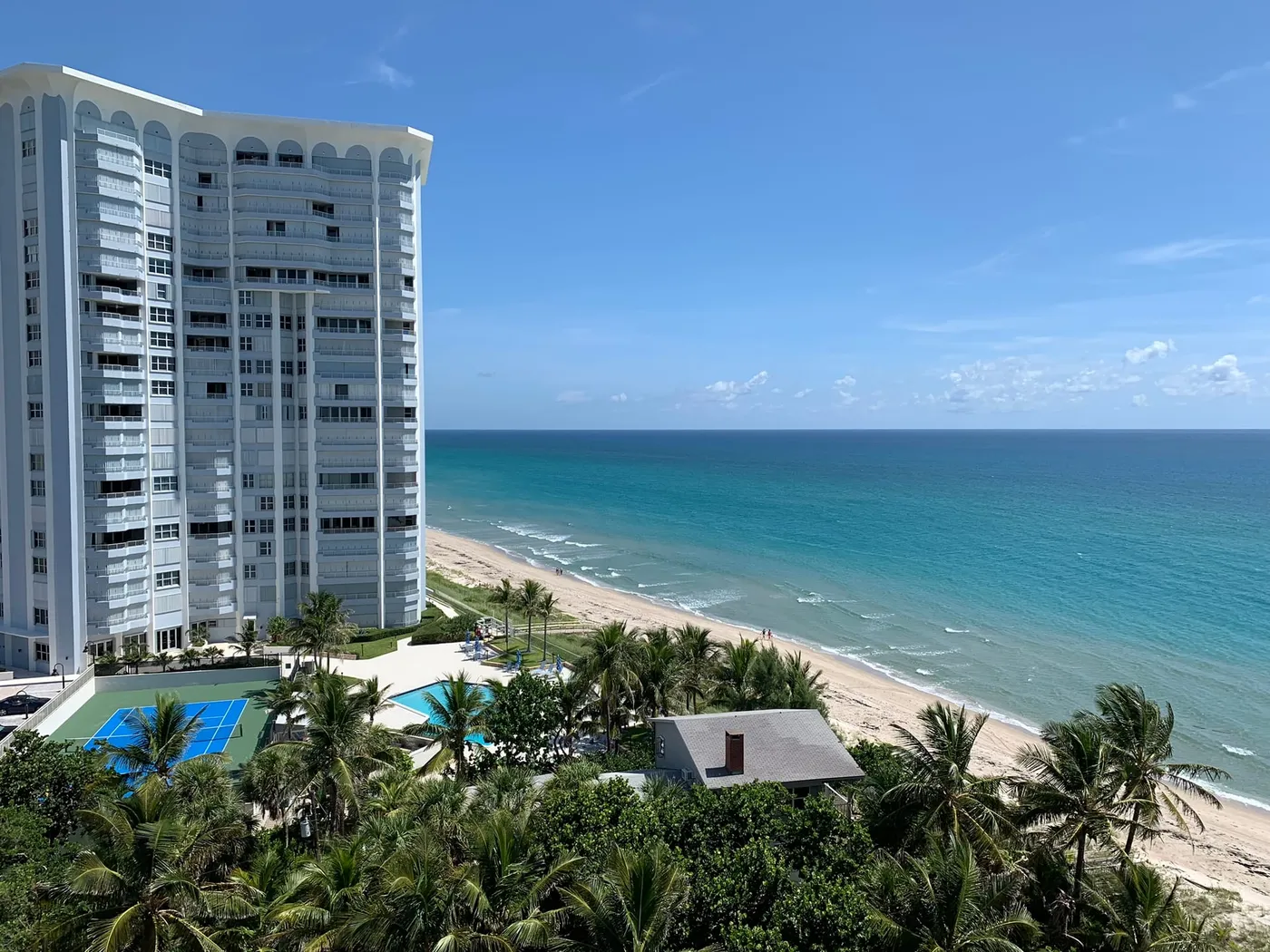 An oceanfront condominium tower seen from above, with its pool and tennis courts beside the beach and the ocean at right