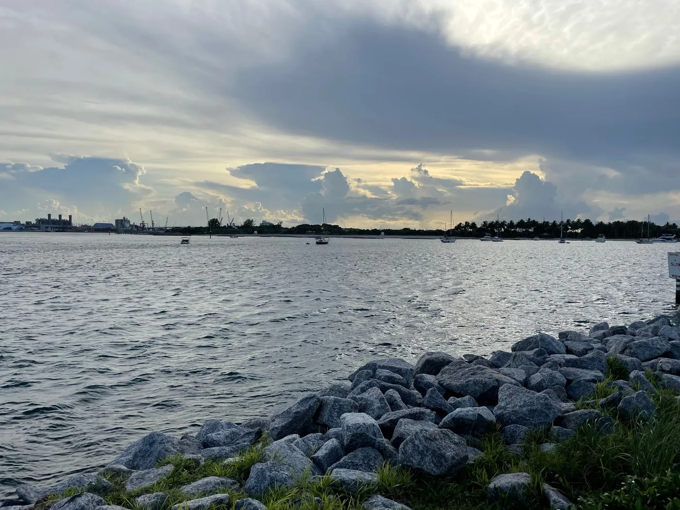 Sailboats at anchor under a darkening sky, with the Port of Palm Beach cranes on the far shore and rocks in the foreground