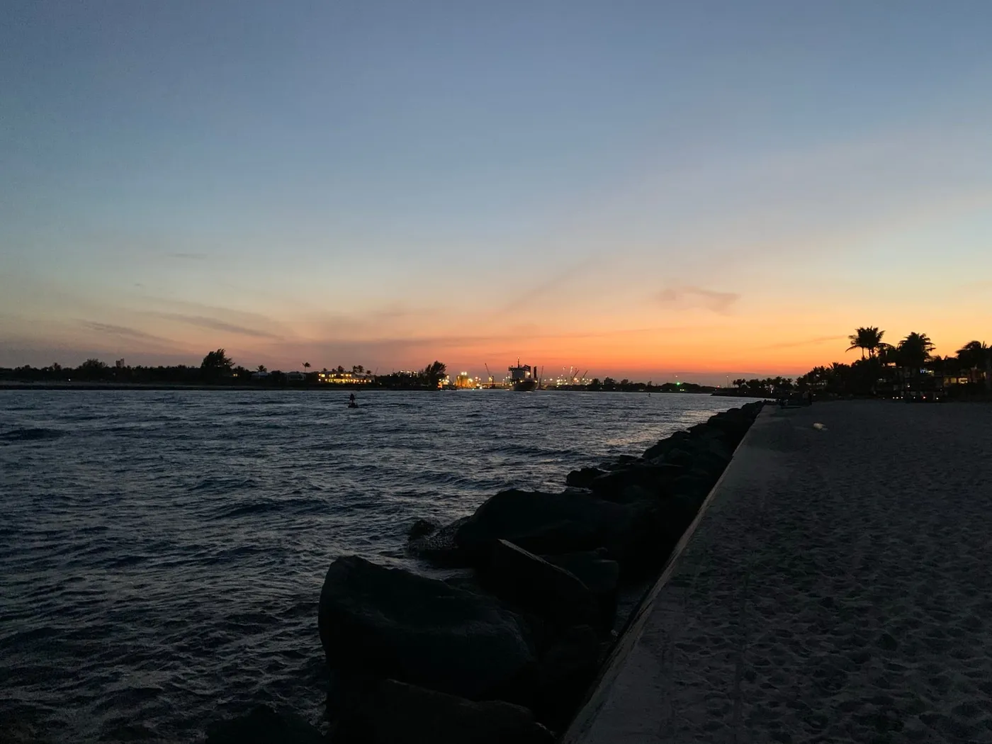 The Lake Worth Inlet at dusk from the sandy shore by the seawall, with the lit Port of Palm Beach cranes on the horizon