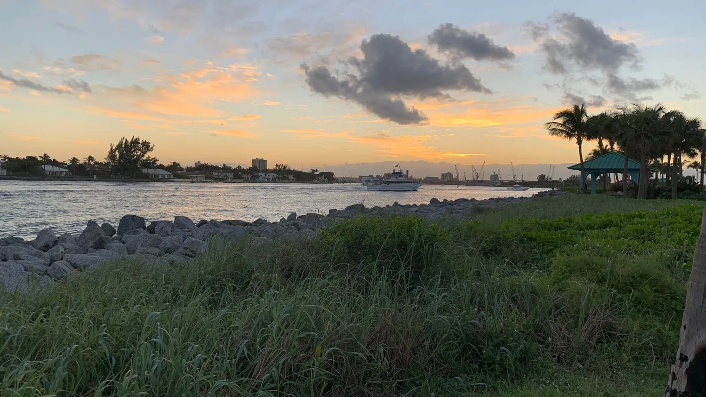 A fishing boat heading in through the Lake Worth Inlet at dusk, with a teal-roofed gazebo and palms at right and port cranes on the horizon