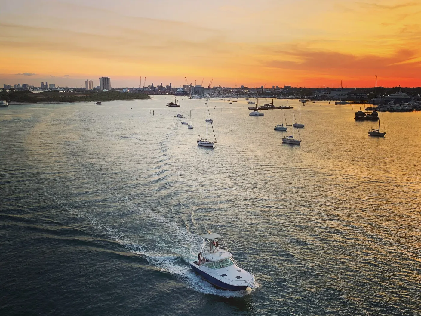 Boats at anchor in the lagoon at sunset, with a sportfishing boat heading out in the foreground and the Port of Palm Beach cranes on the horizon