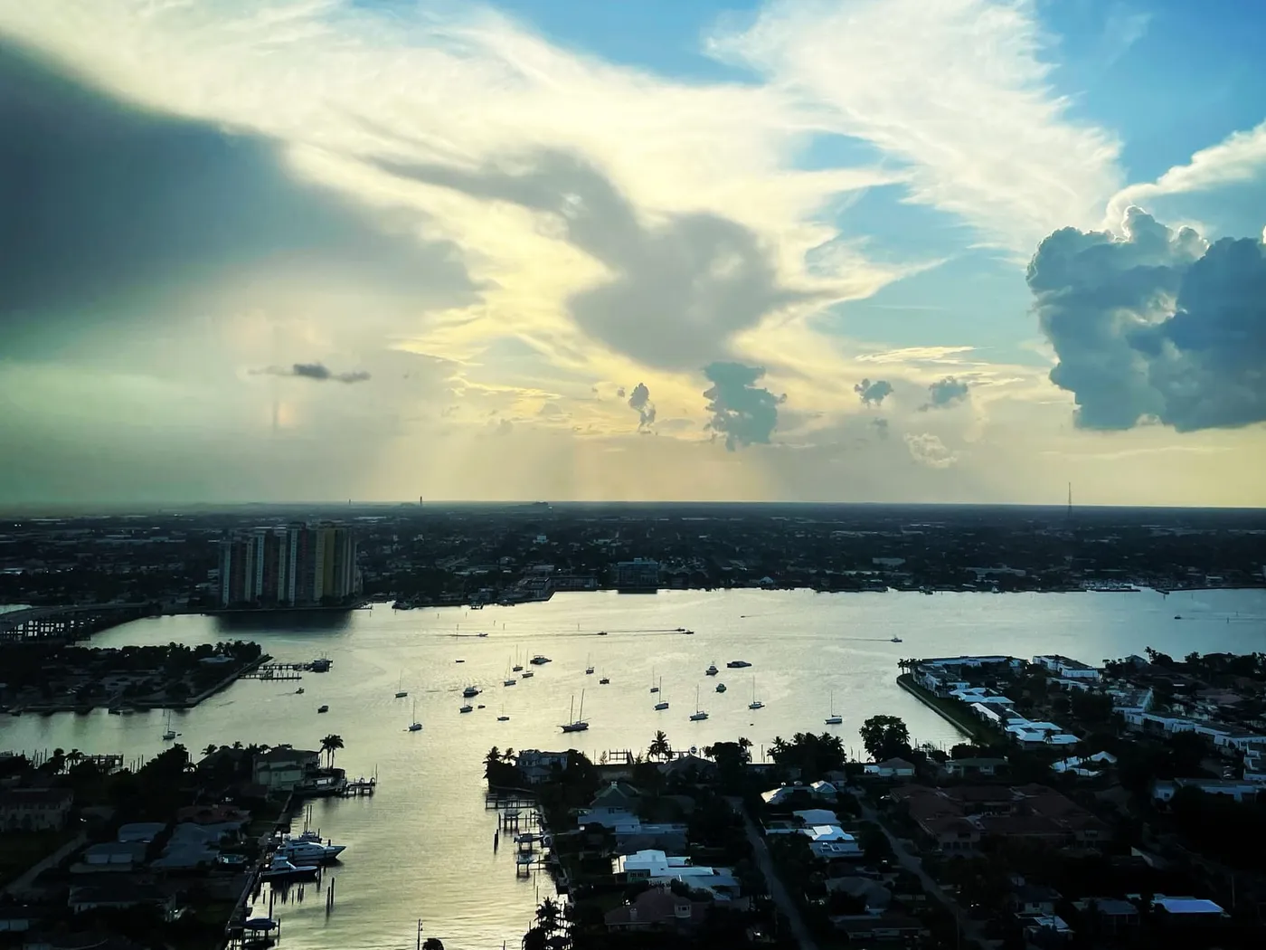 A high view west over lagoon-side house streets, canals and boats at anchor on the lagoon, under a dramatic evening sky