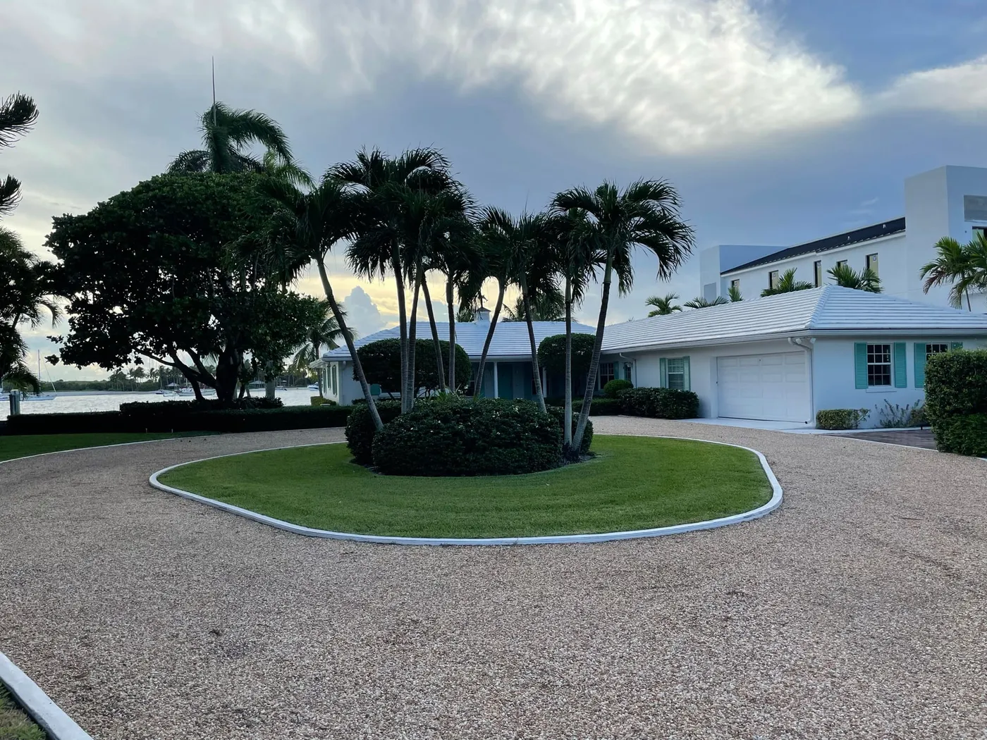 A white single-story house with a gravel circular drive and palms, with the lagoon and boats at anchor visible beyond