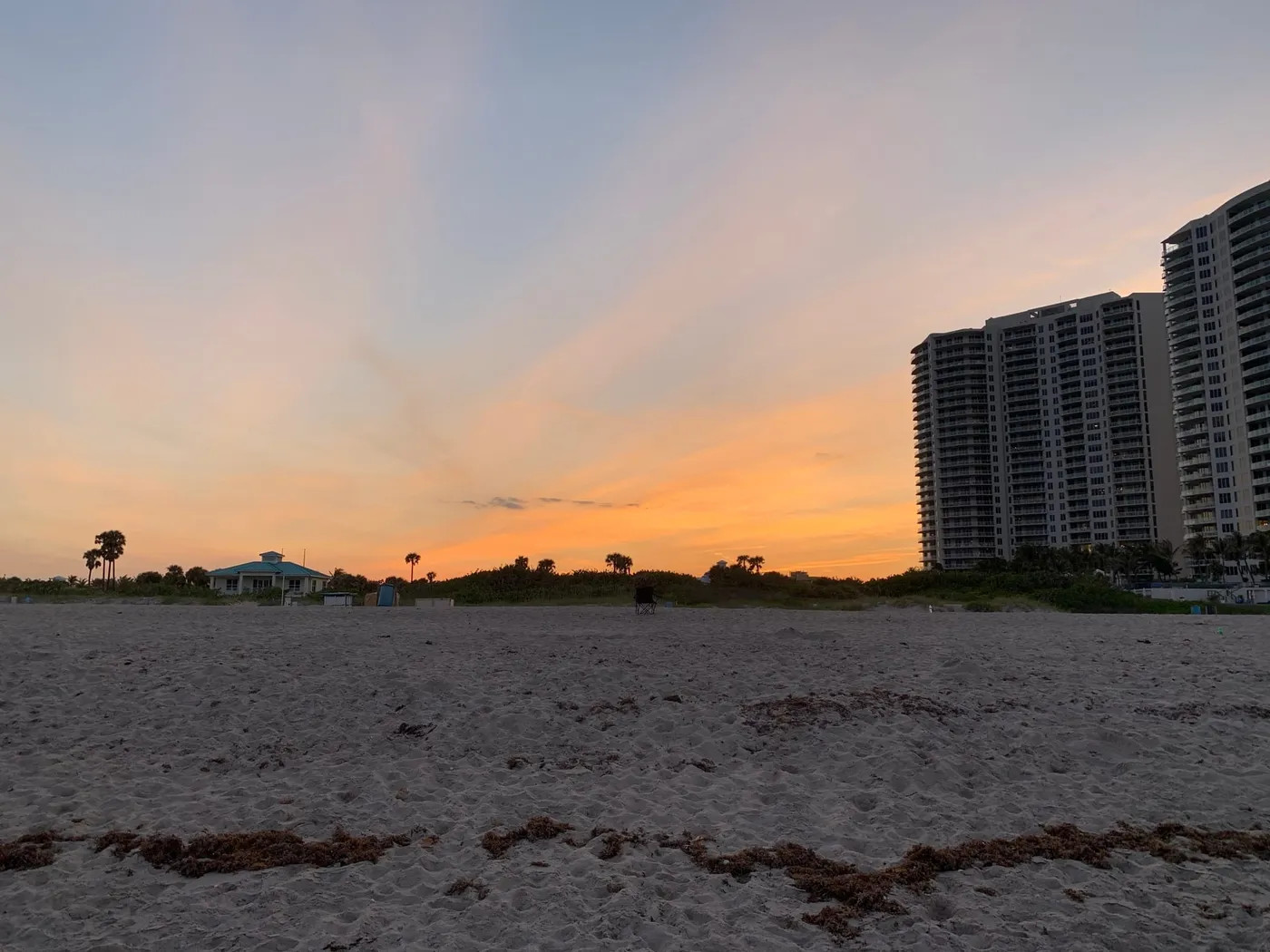 A public beach at dusk under an orange sky, with a teal-roofed pavilion at left and condominium towers at right
