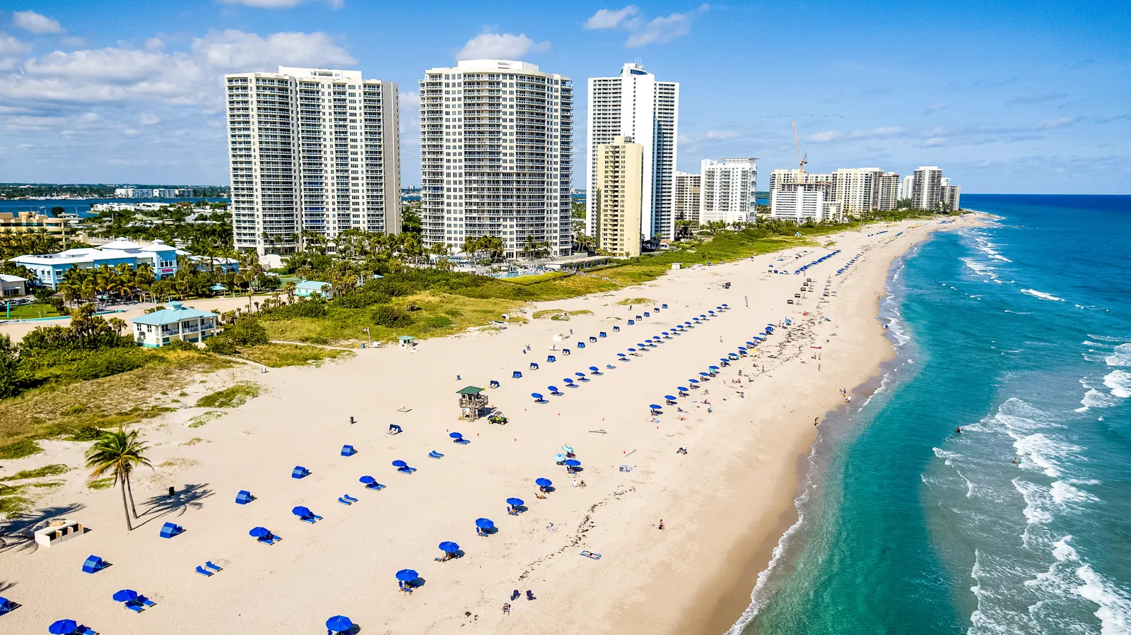 Aerial view of a wide white-sand beach with blue umbrellas and high-rise condo towers along the Atlantic shoreline
