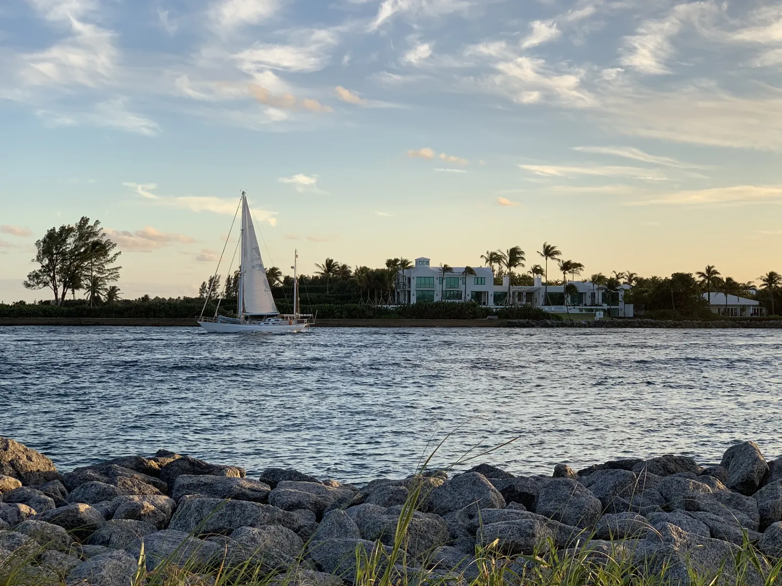 Sailboat passing a modern waterfront estate at golden hour