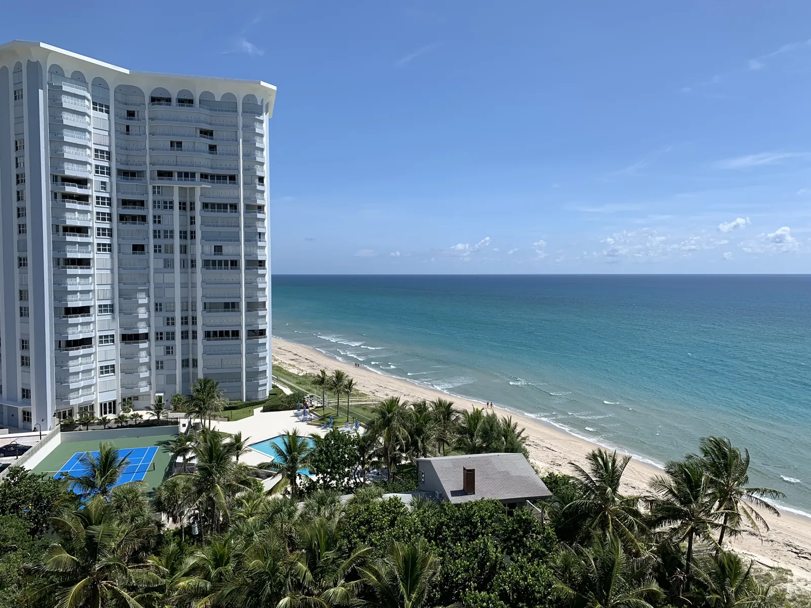 Elevated oceanfront view of a white high-rise condo tower, pool, tennis court, palms, and Atlantic beach