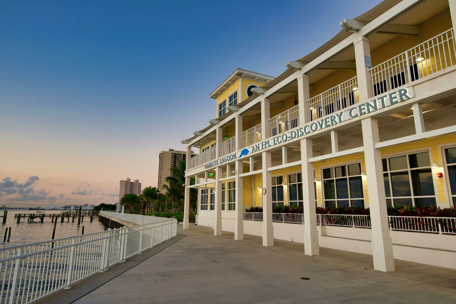 Yellow two-story waterfront eco-discovery center at dusk with white columns and railed boardwalk
