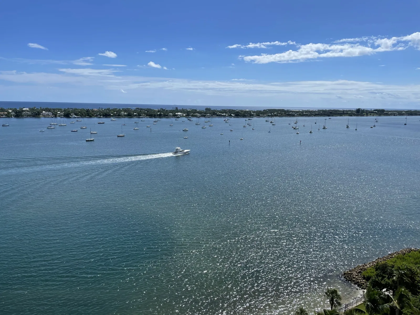 Elevated view over a sparkling Intracoastal-style waterway with moored sailboats, a motor yacht leaving a wake, and a low barrier island with homes on the far shore