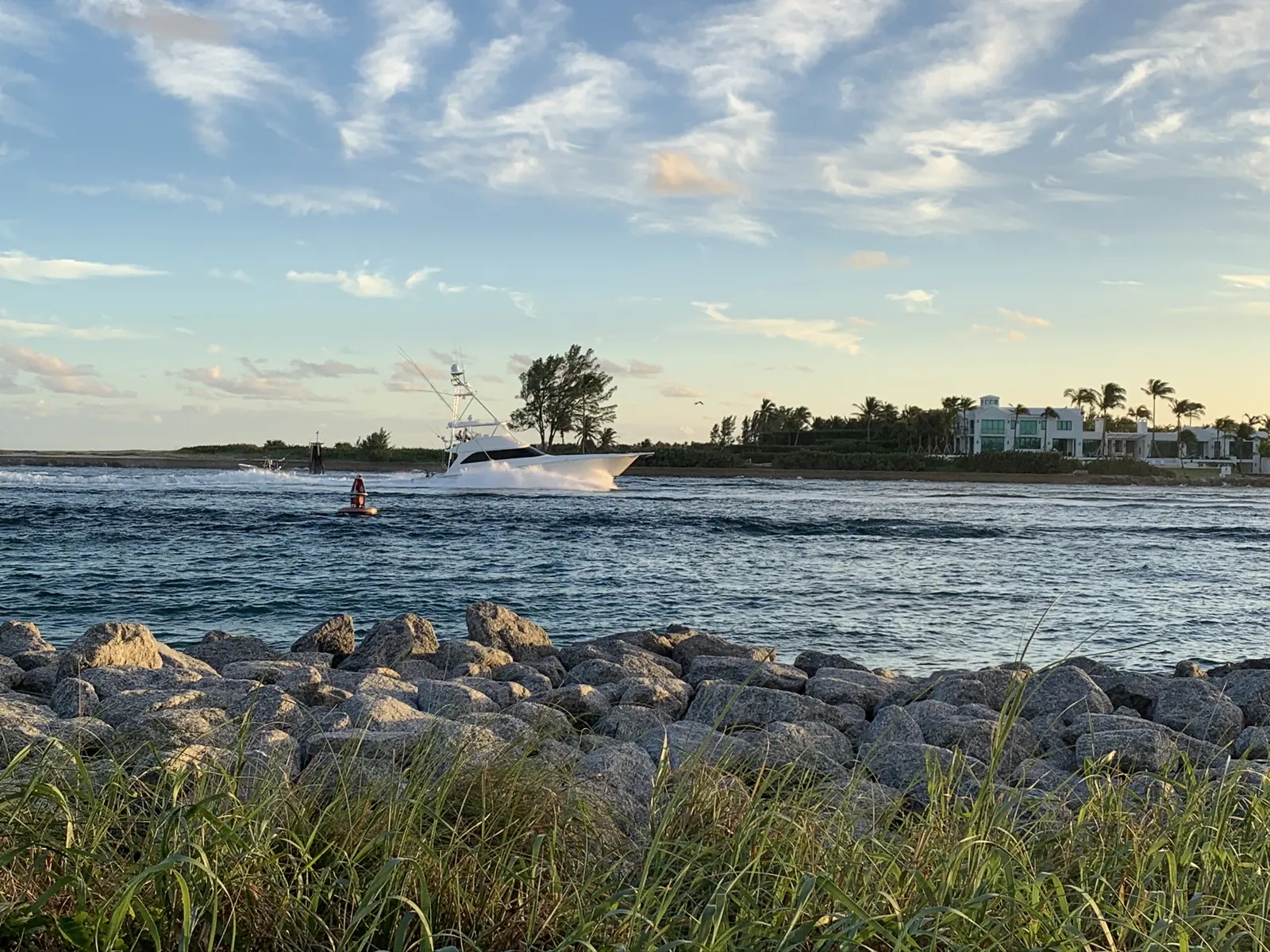 Sportfishing yacht running an inlet past a rock jetty at golden hour, with a modern waterfront estate on the far shore