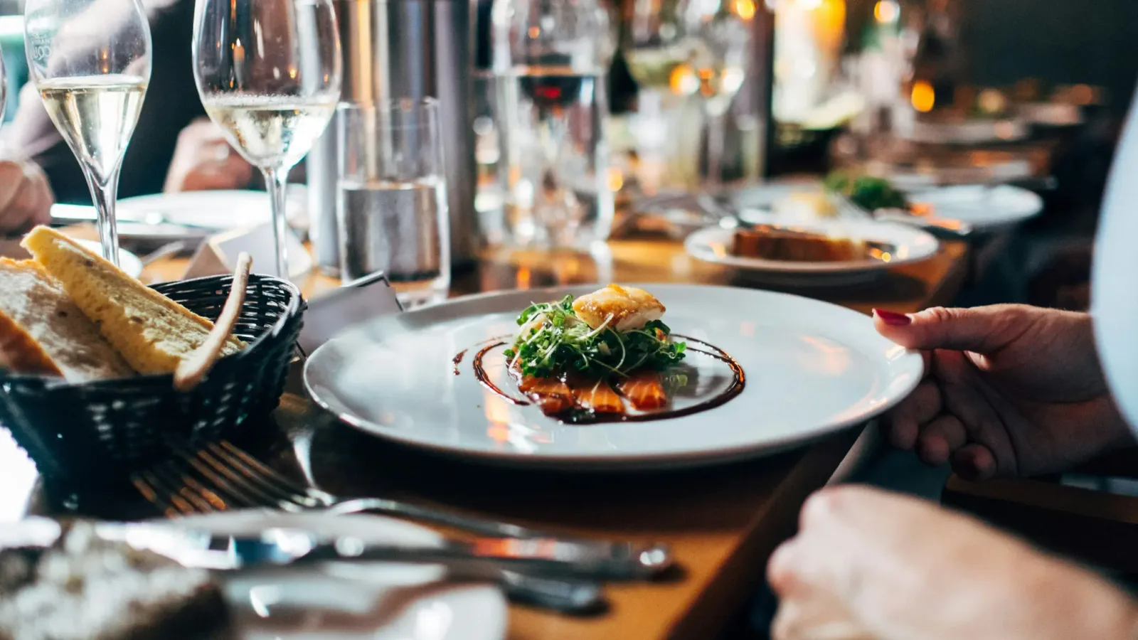 A plated fish course with microgreens beside bread and wine glasses on a set restaurant table