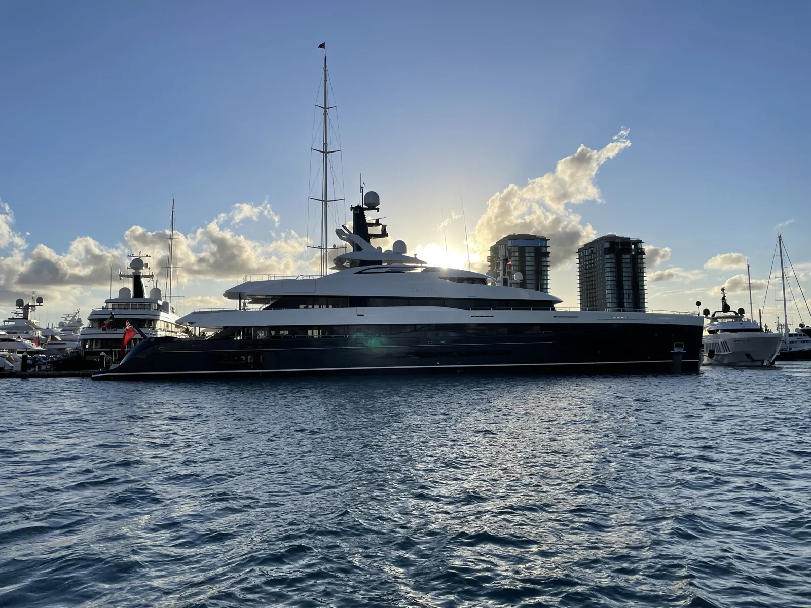 Large dark-hulled superyacht docked in a marina at sunset with high-rise towers behind