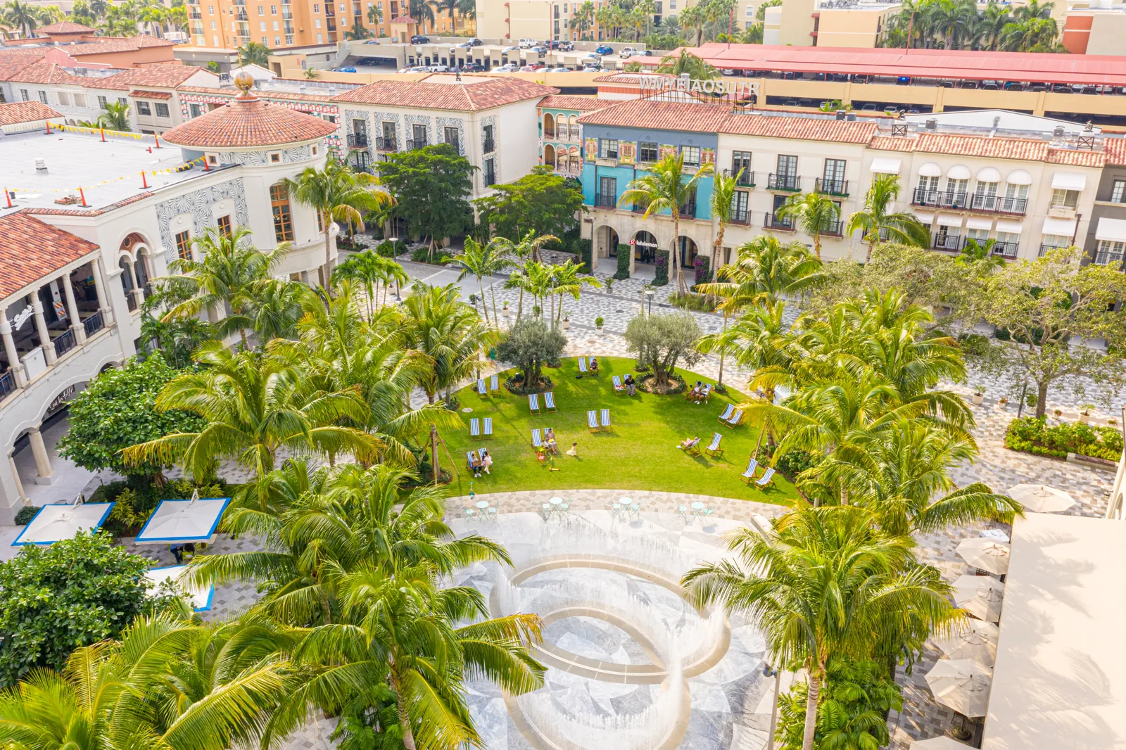 Elevated view of a Mediterranean-style outdoor shopping plaza with lawn, fountain, and palm trees