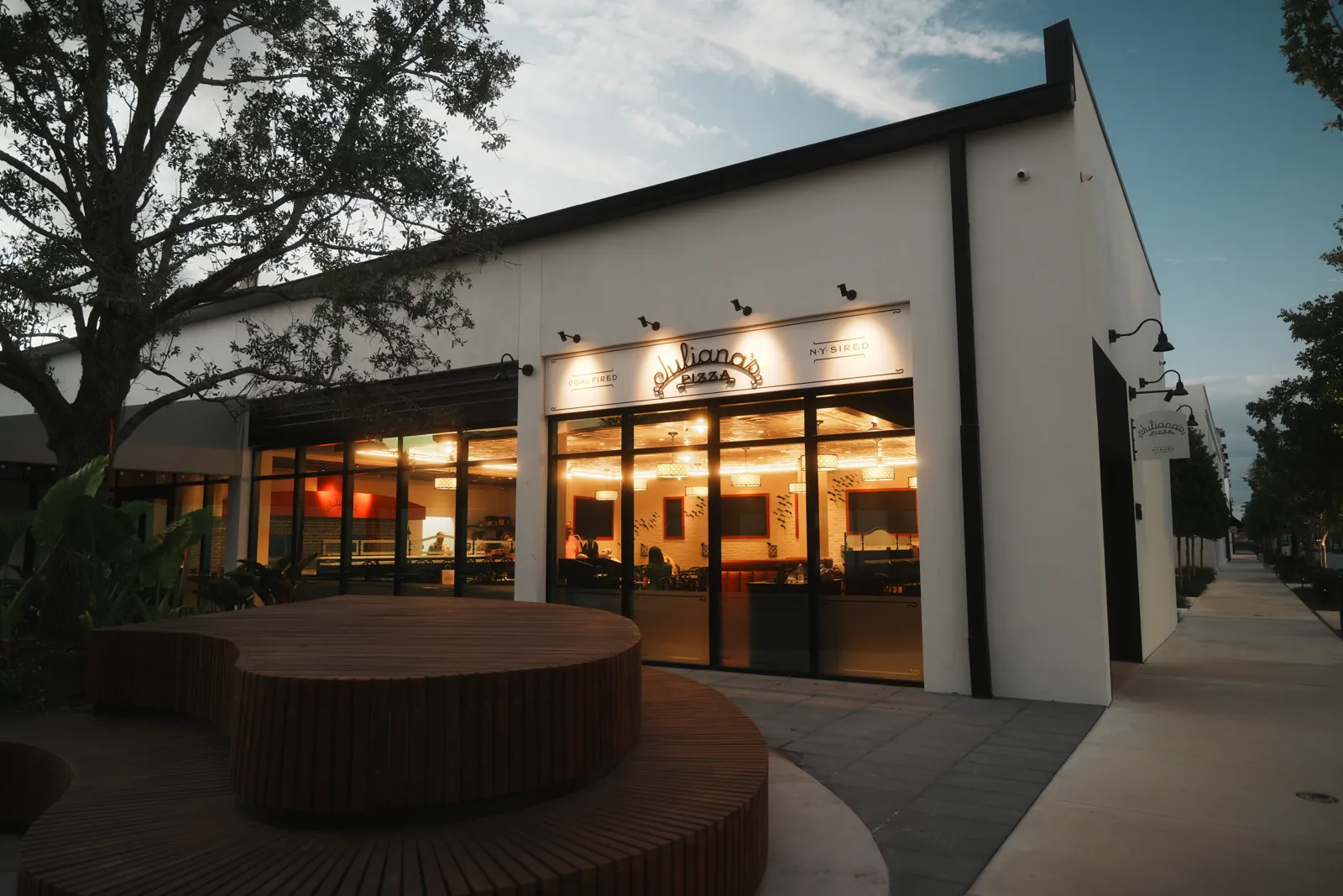 Pizza restaurant storefront at dusk with warm glowing interior and curved wooden bench in foreground