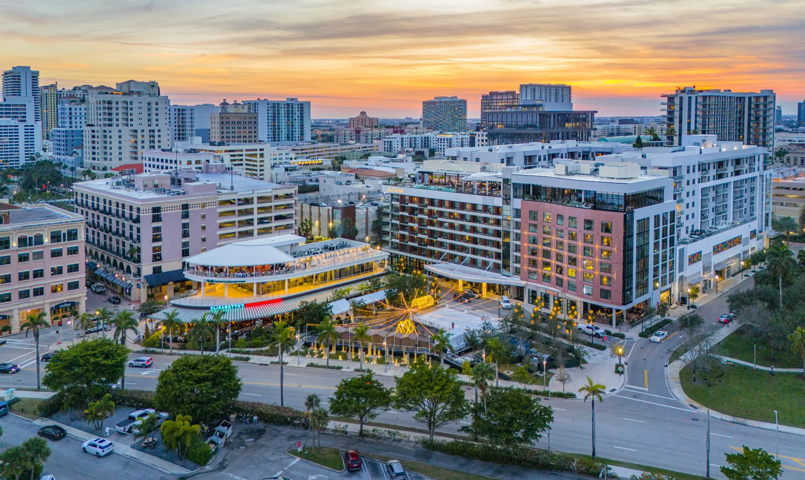 Aerial dusk view of a mixed-use downtown district with a lit open-air dining pavilion, string-lit plaza, and mid-rise hotel/residential buildings