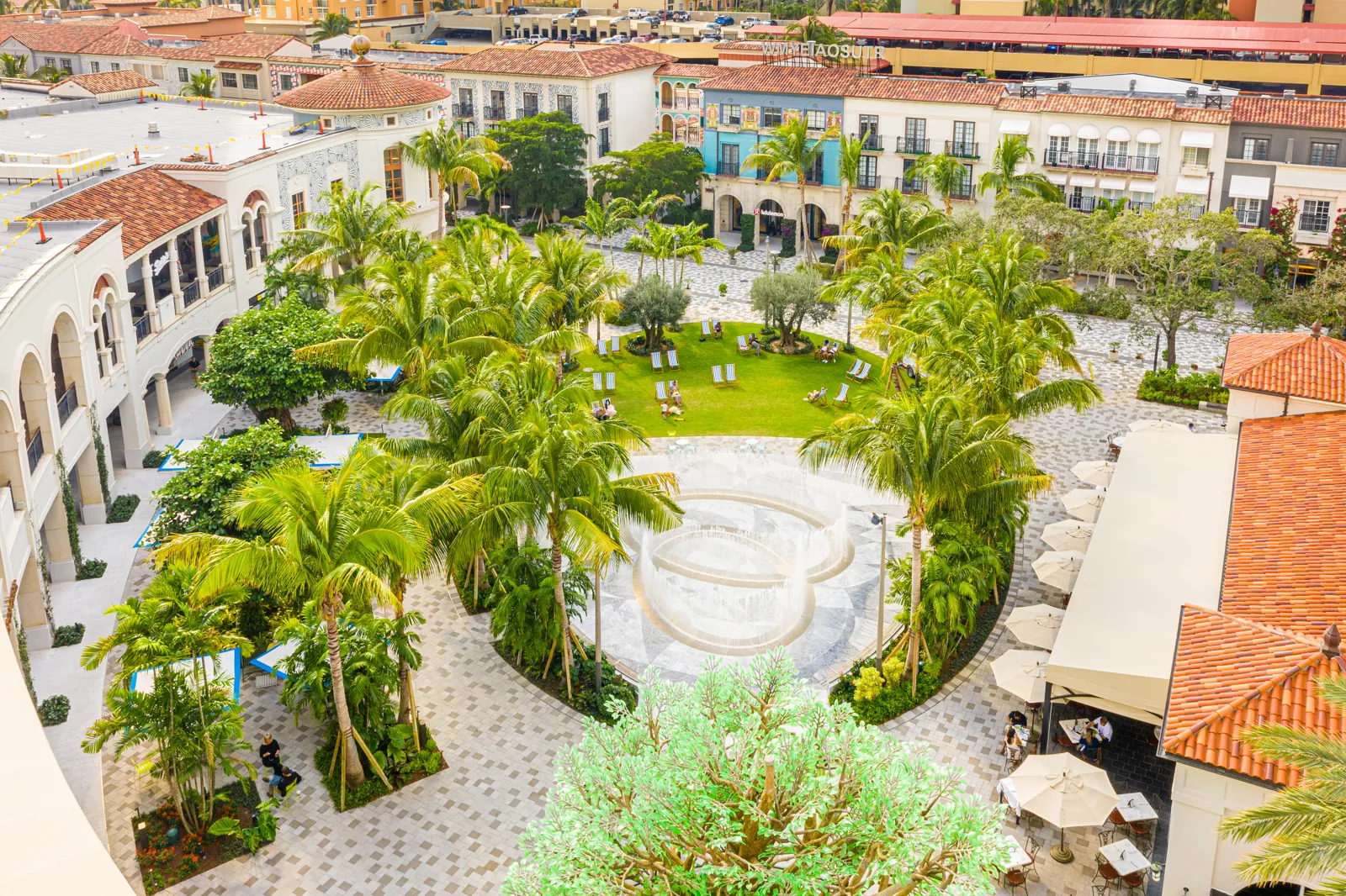Elevated view of a Mediterranean-style outdoor shopping plaza with central lawn, fountain, and palm-lined courtyard