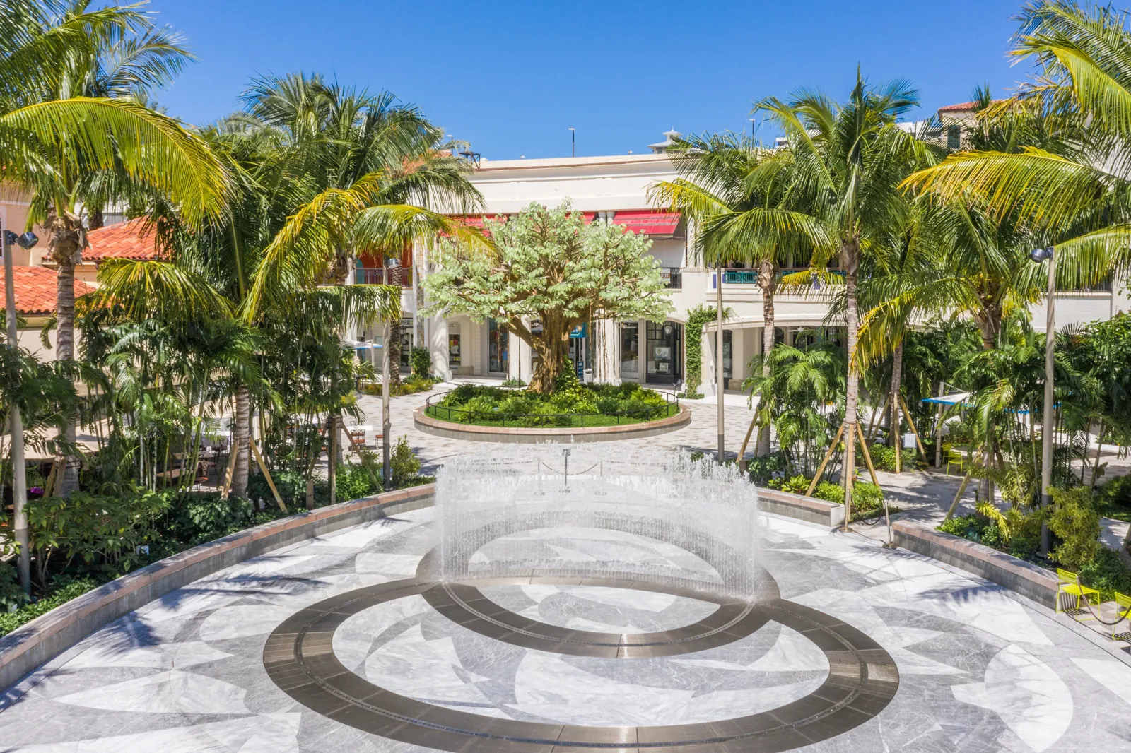 Open-air shopping plaza courtyard with marble fountain plaza, palm trees, and sculptural tree centerpiece