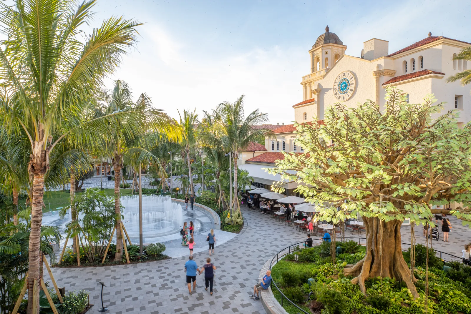 Open-air Mediterranean Revival plaza with interactive fountain, clock tower building, and outdoor cafe seating at golden hour
