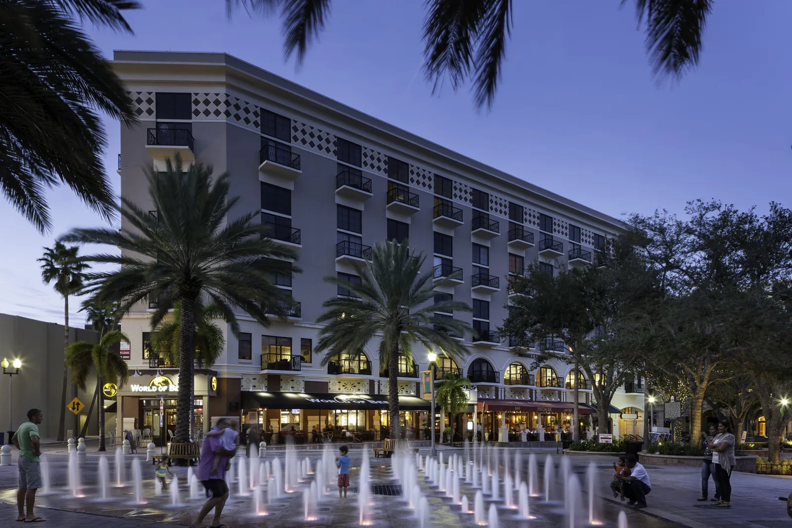Mixed-use plaza at dusk with interactive fountain, palm trees, and a multi-story building with ground-floor restaurants