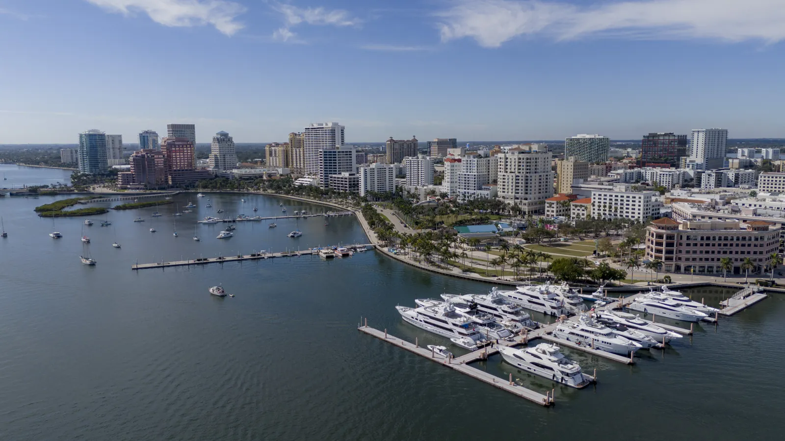 Aerial view of a waterfront downtown skyline with a superyacht marina and palm-lined waterfront park