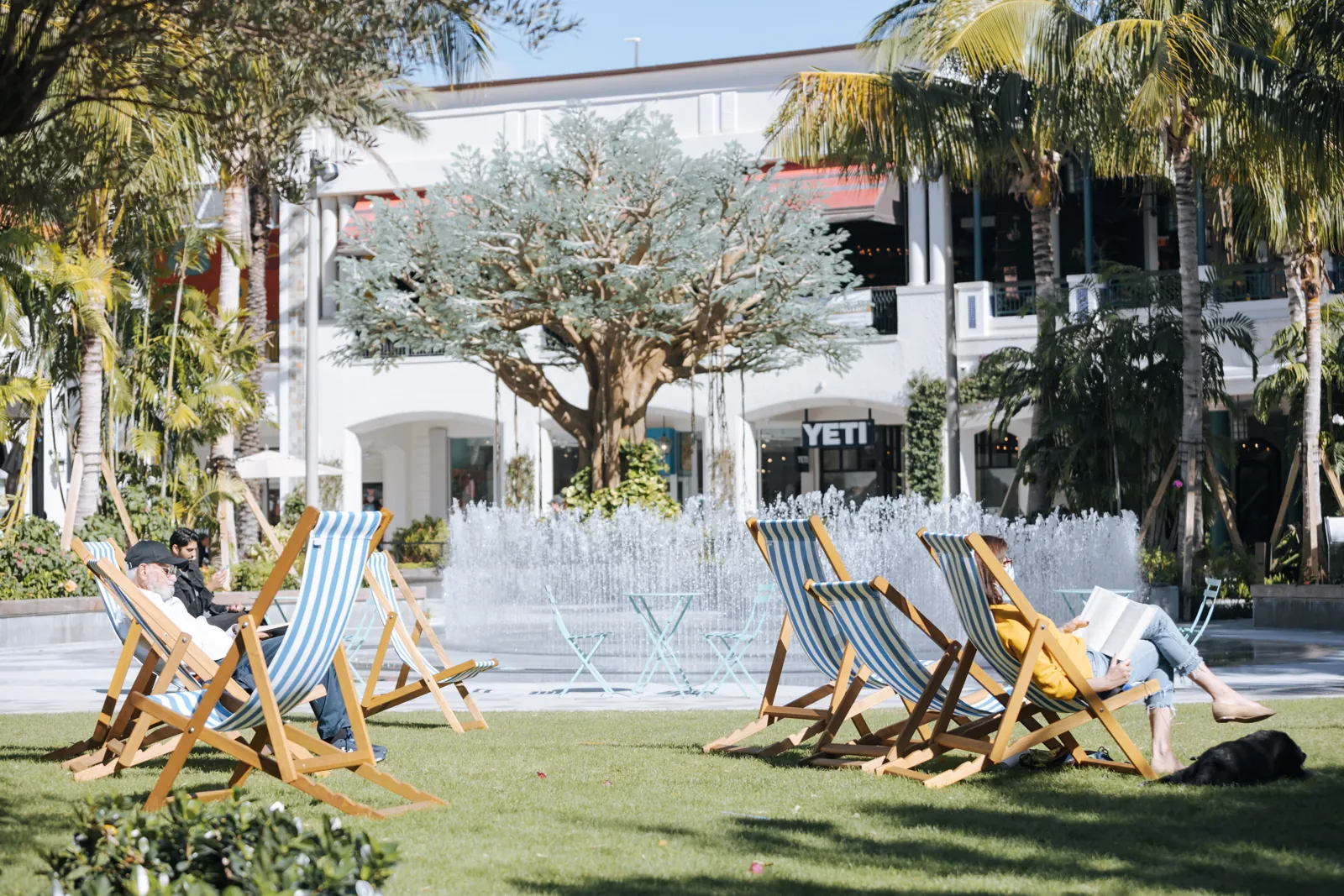 Striped deck chairs on a sunny lawn facing a splash-pad fountain at an outdoor shopping plaza