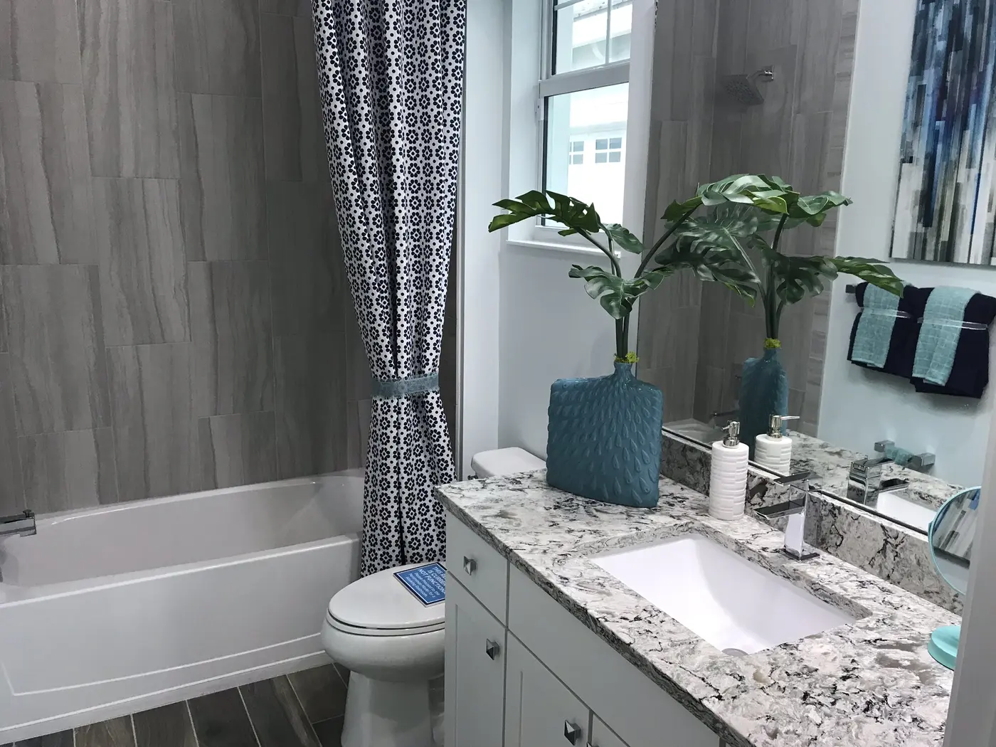 A bathroom with a tub and patterned shower curtain, a granite vanity and a potted plant in a Minto model home in Westlake