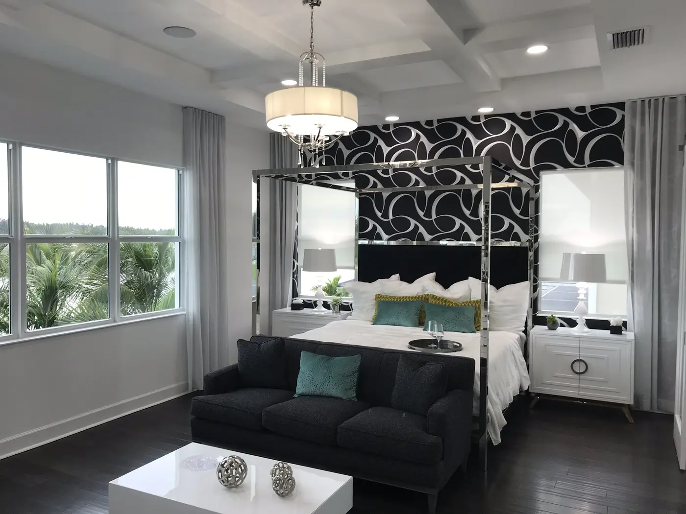 A bedroom with a four-poster metal canopy bed, a black patterned accent wall, a dark sofa and windows on two sides in a Minto model home in Westlake