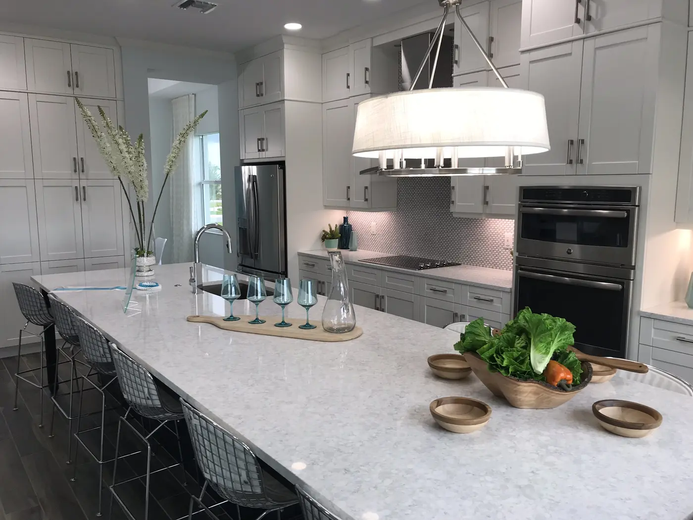 A white kitchen with a long quartz island, metal bar stools, a drum pendant light and stainless wall ovens in a Minto model home in Westlake