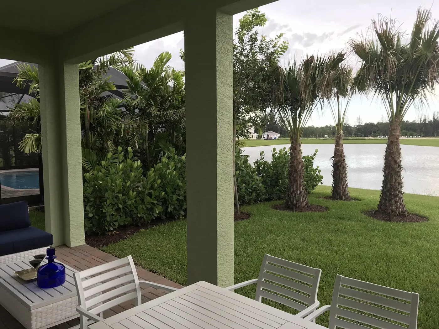A covered patio with white outdoor chairs at a Minto model home in Westlake, looking onto a lawn with three palms, shrubs and a lake