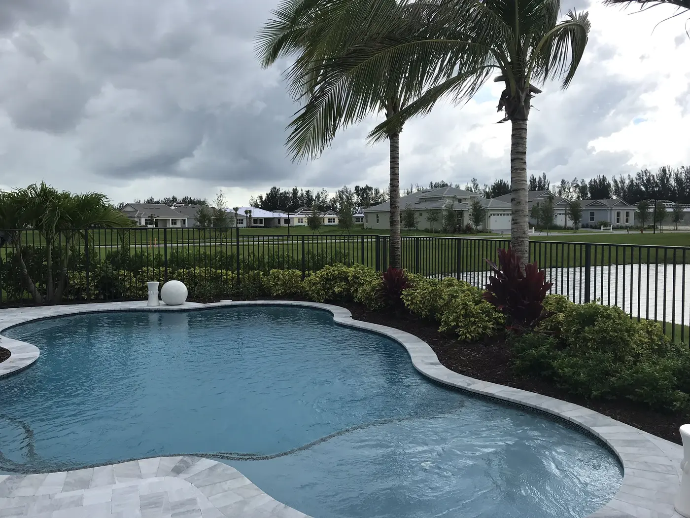 A curving pool with a stone ball fountain behind a black metal fence at a Minto model home in Westlake, with a lake and a row of single-story homes beyond under a cloudy sky
